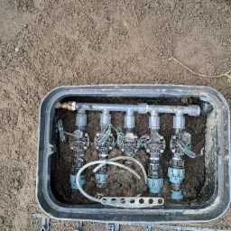 Czarny plastikowy zawór nawadniający z pięcioma zaworami elektromagnetycznymi, częściowo zakopany w ziemi obok gałązki iglaka.