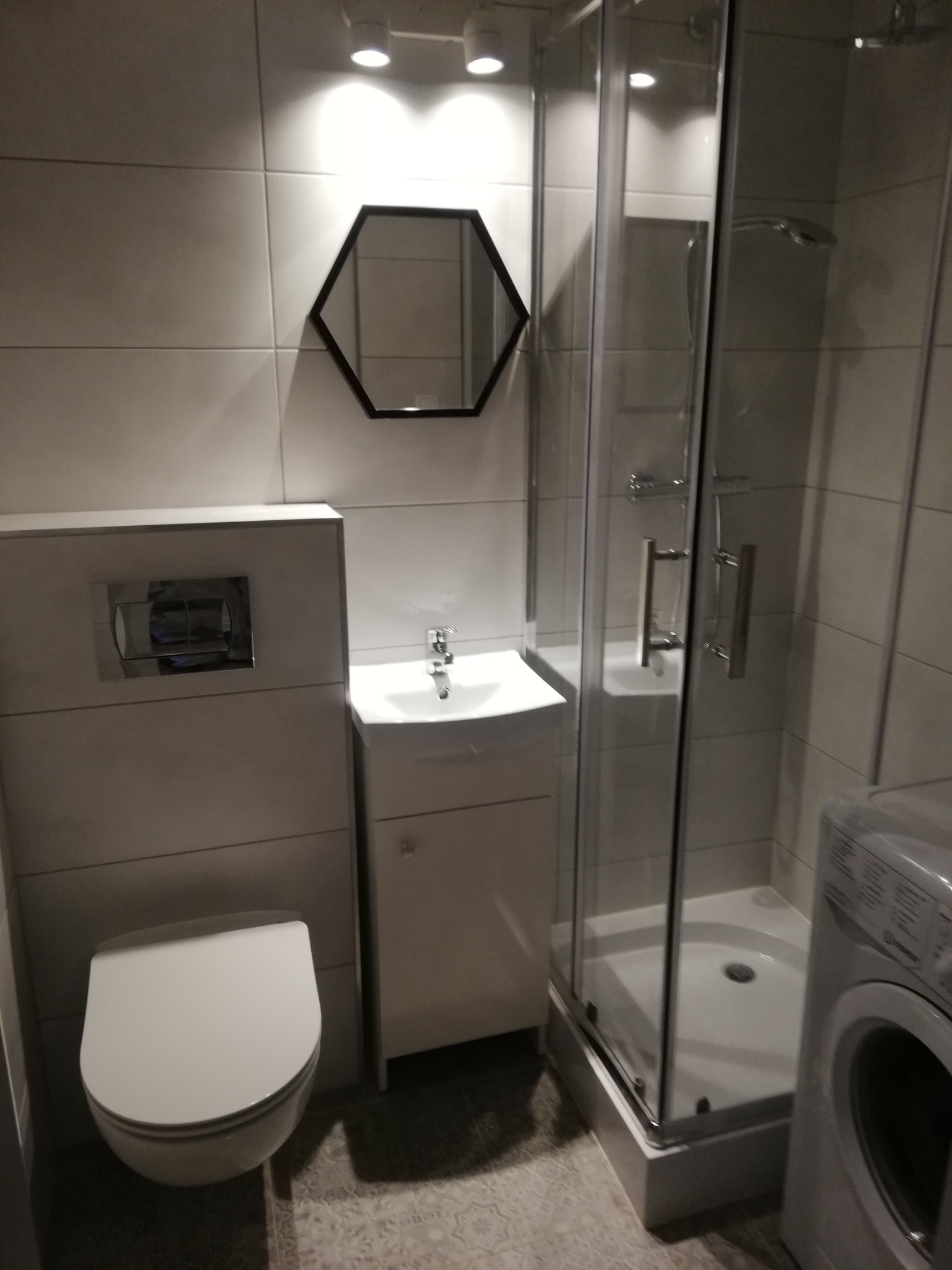 Nowoczesna łazienka z minimalistycznym wystrojem: białe płytki, hexagonalne lustro, kabina prysznicowa i pralka.