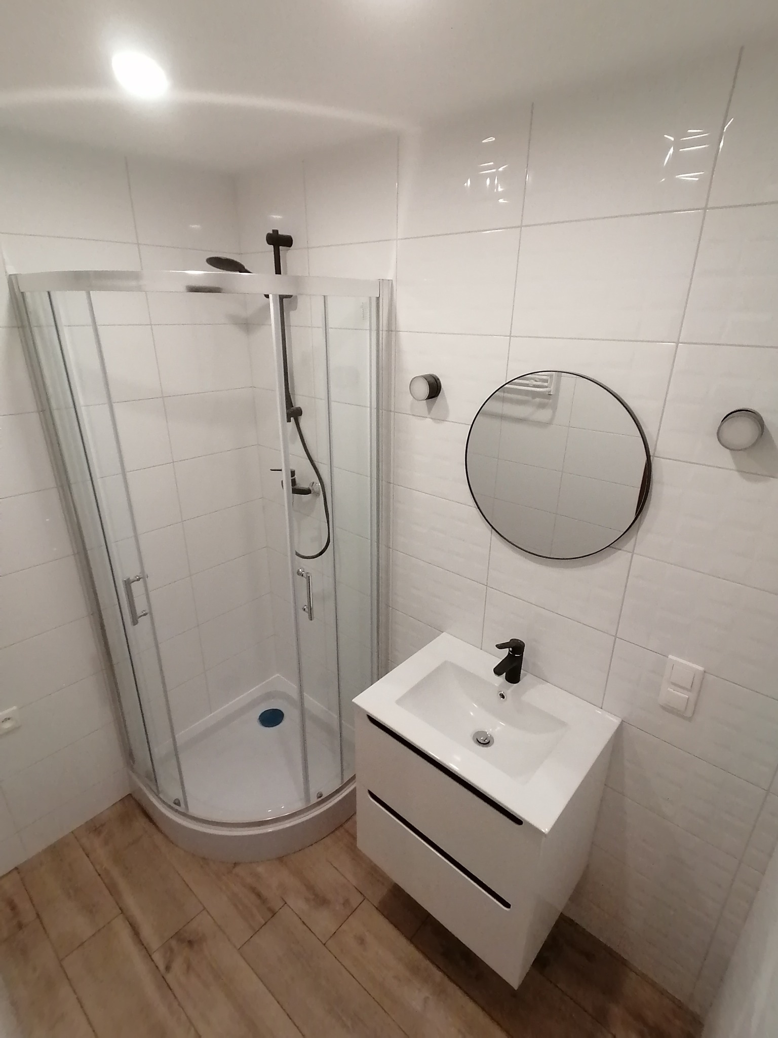 Nowoczesna łazienka z kabiną prysznicową z zaokrąglonymi drzwiami, białą umywalką z czarną baterią, okrągłym lustrem w czarnej ramie i drewnopodobną podłogą.