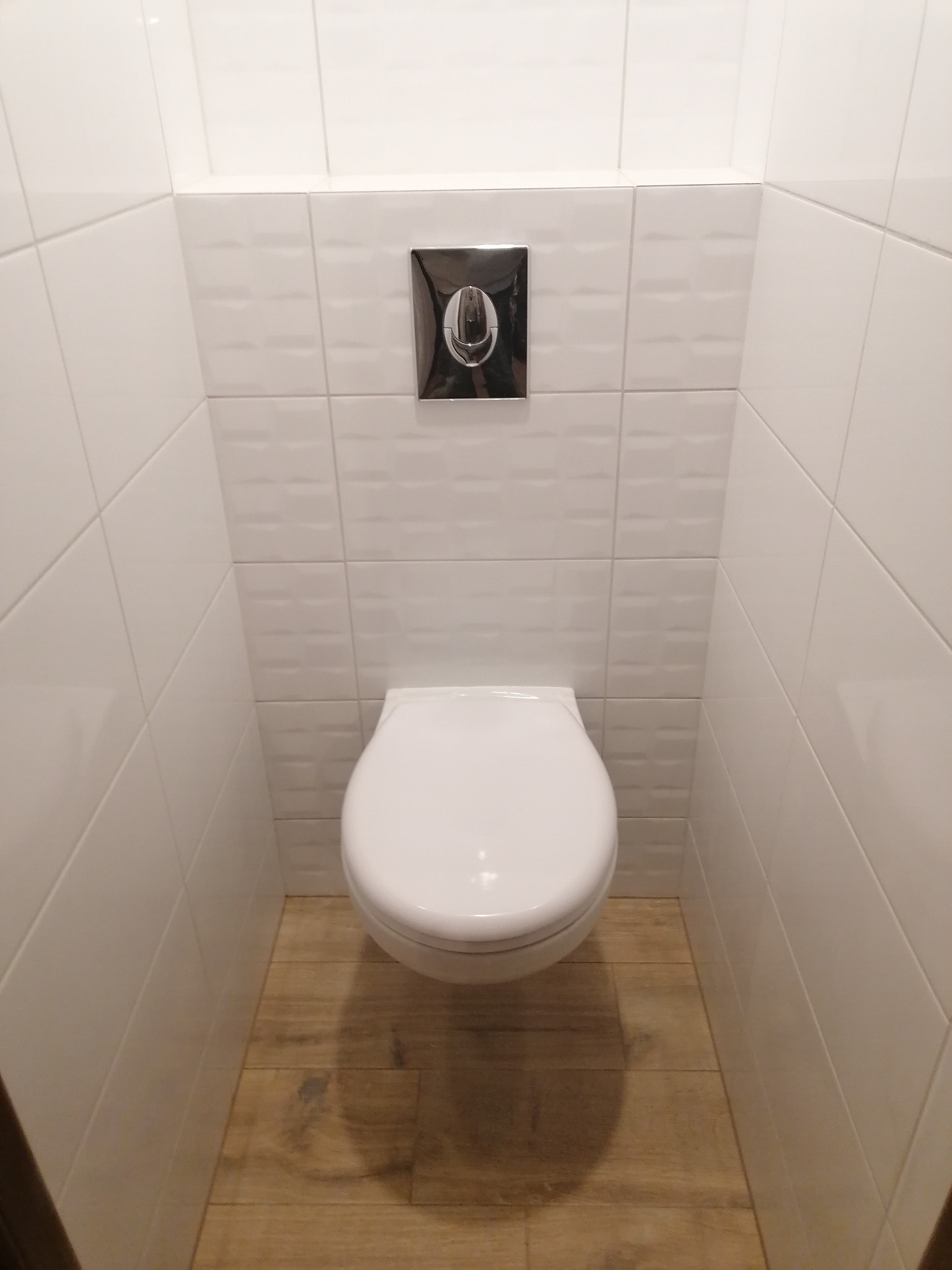 Kompaktowa toaleta podwieszana z chromowanym przyciskiem spłukującym, wykończona białymi płytkami ściennymi i podłogą imitującą drewno, w łazience w Kórniku.