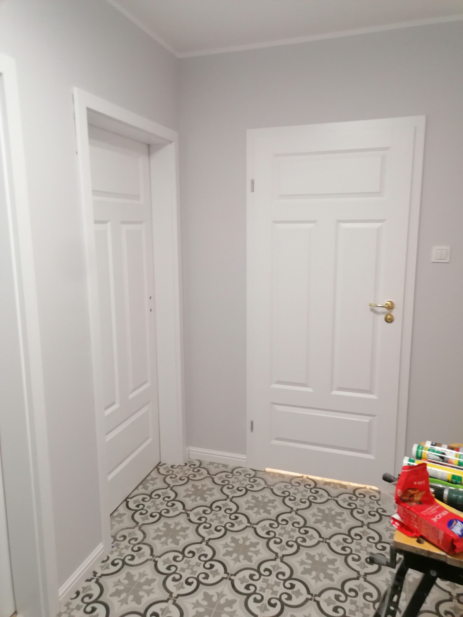 Wykończony korytarz z szaro-białymi ścianami, dwoma białymi drzwiami z frezowanymi panelami i wzorzystą szaro-białą podłogą z płytek, widoczne materiały budowlane na stojaku.