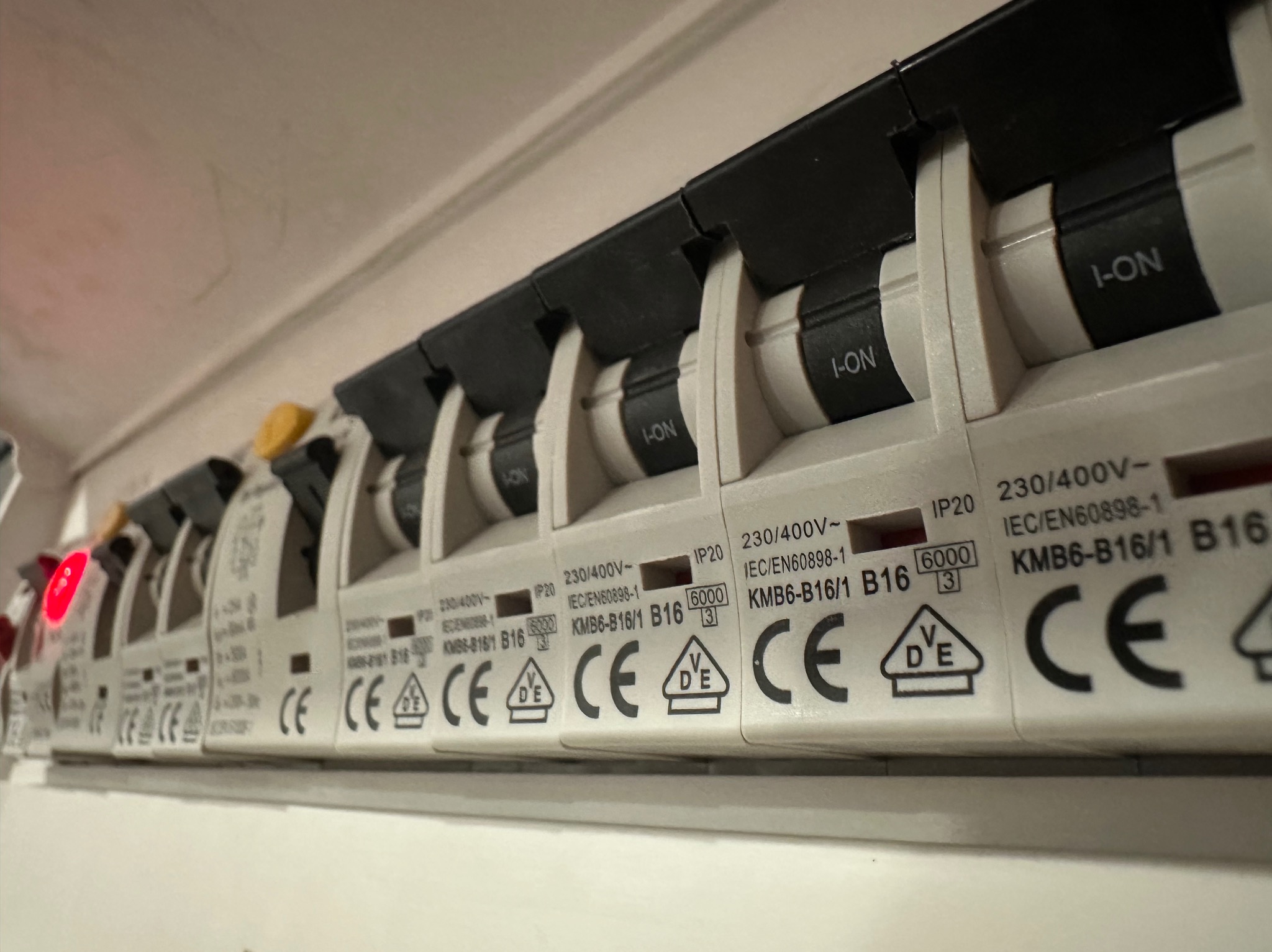 Rząd wyłączników nadprądowych B16 w rozdzielnicy, jeden z nich z włączonym obwodem sygnalizowanym czerwoną diodą, widoczne oznaczenia parametrów i certyfikaty na obudowach.