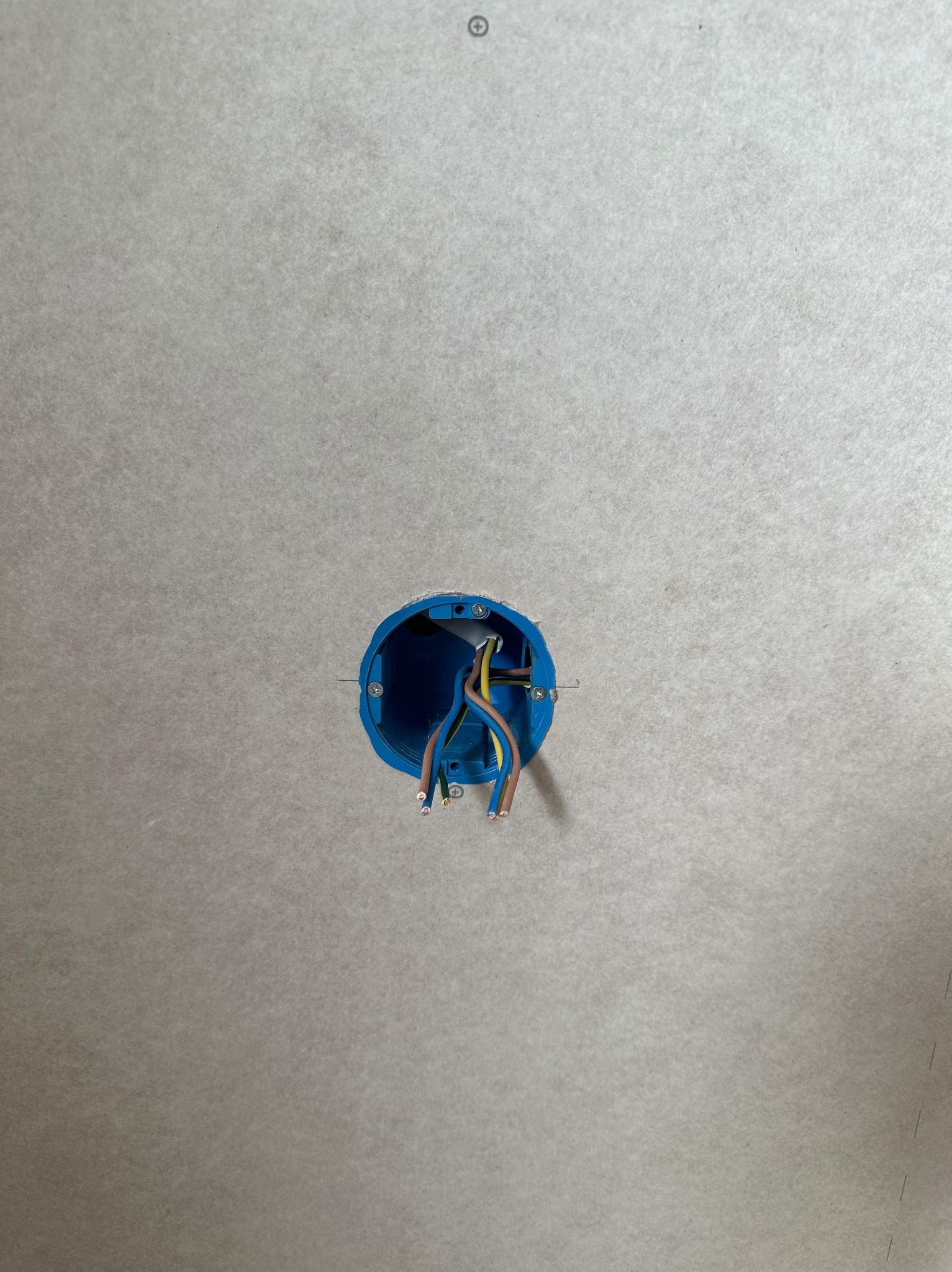 Niebieska puszka elektryczna zamontowana w płycie gipsowo-kartonowej z wystającymi przewodami o różnych kolorach izolacji.