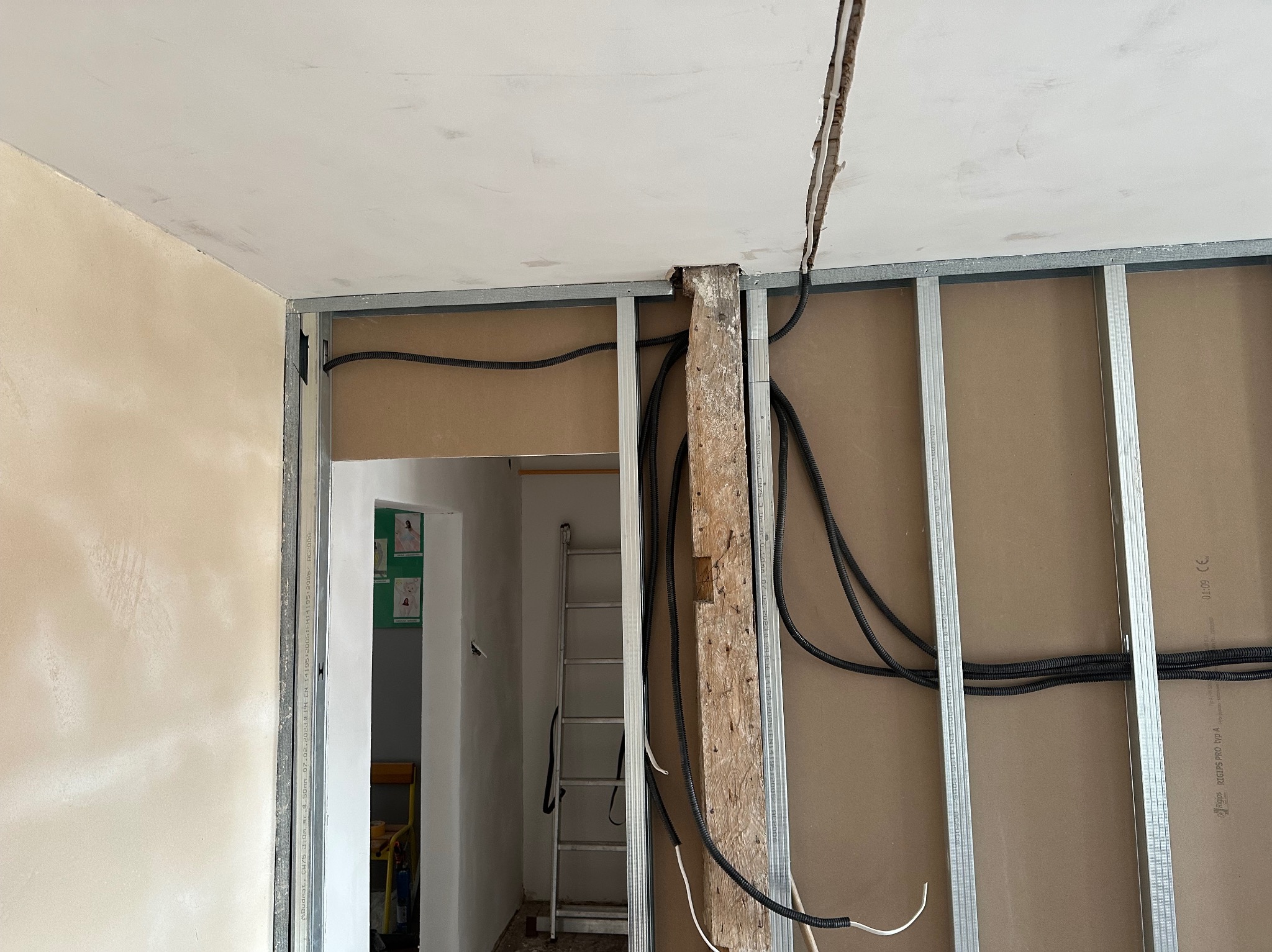 Montaż instalacji elektrycznej wewnątrz ściany z płyt kartonowo-gipsowych na metalowym stelażu, widoczne kable w peszlach oraz drewniany element wspierający konstrukcję, w tle drabina.