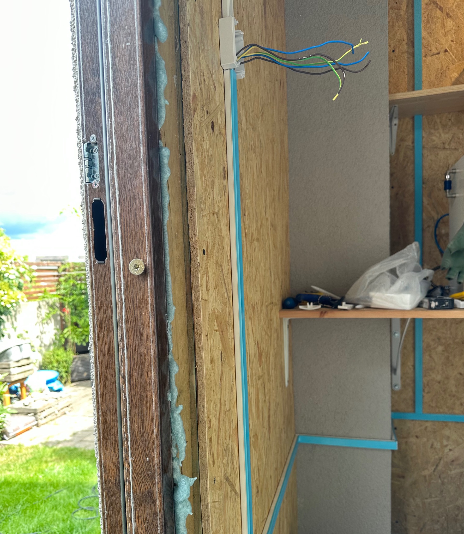 Wnętrze budynku w trakcie wykańczania: widoczne przewody elektryczne wychodzące z puszki, ocieplona rama okienna pianką montażową, płyty OSB z niebieskimi profilami i półka z narzędziami w tle.