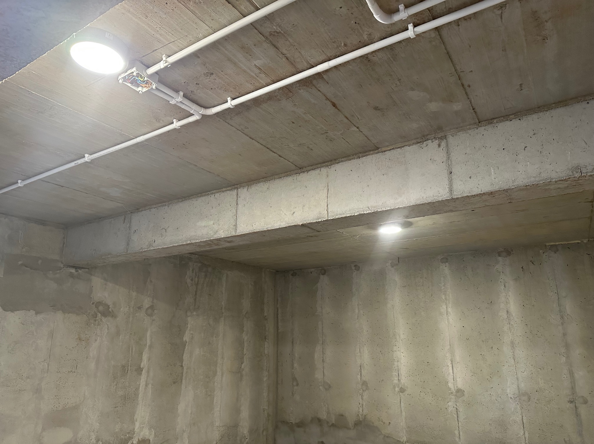 Surowy betonowy sufit z widocznymi przewodami elektrycznymi w białych rurkach, przymocowanymi plastikowymi uchwytami, oraz puszką instalacyjną i zamontowanym okrągłym oświetleniem LED.