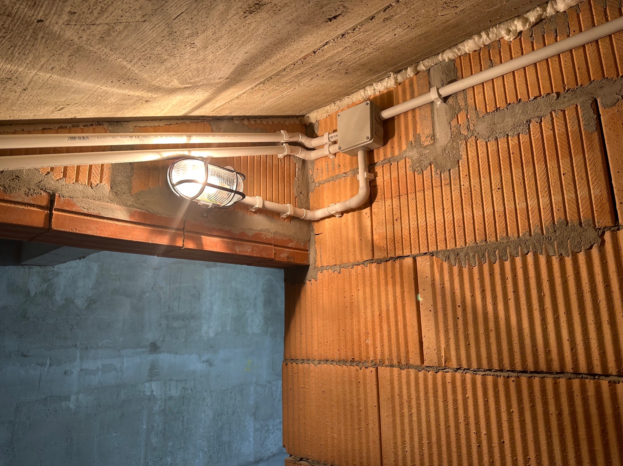 Lampa sufitowa w metalowej klatce ochronnej, zamontowana na ścianie z cegły dziurawki w surowym stanie, z widocznymi białymi rurami PCV prowadzącymi do szarej puszki elektrycznej, pianka montażowa...