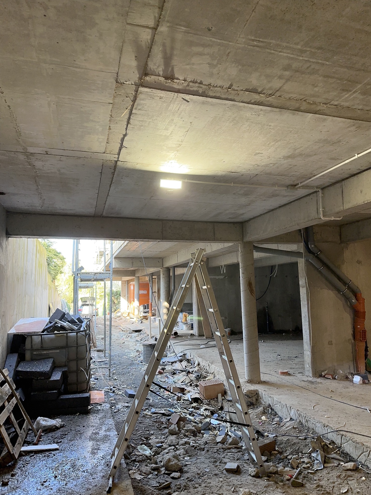Wnętrze w stanie surowym z widoczną drabiną, gruzem i prowizorycznym oświetleniem pod stropem. Widoczne rury instalacyjne przy ścianie i przewody elektryczne pod sufitem.