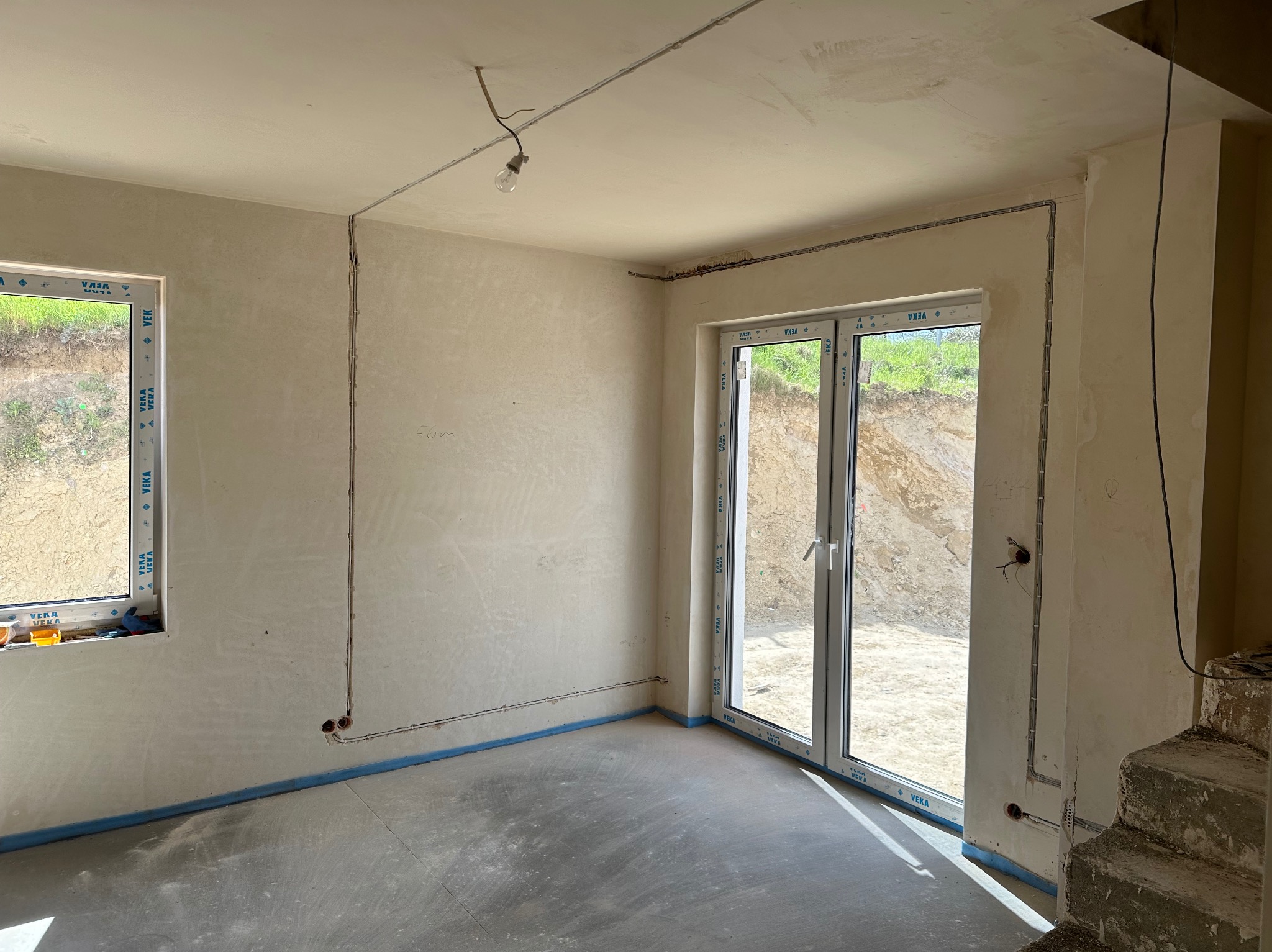 Surowe wnętrze w trakcie remontu z widocznymi przewodami elektrycznymi poprowadzonymi po ścianach, sufitowe gniazdo żarówki z wiszącym kablem, surowy betonowy podkład, okno i drzwi balkonowe...