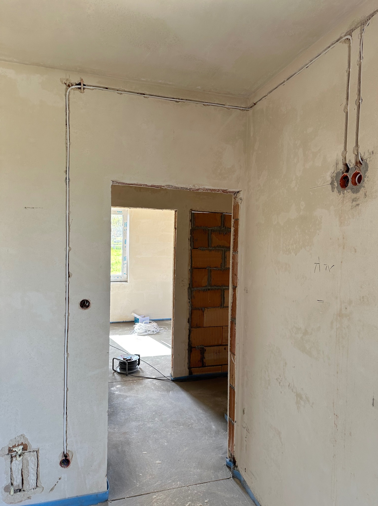 Wnętrze w trakcie remontu: ściany w gładzi, widoczne kable elektryczne w bruzdach, puszki instalacyjne, przewód elektryczny na szpuli w tle, surowy beton na podłodze, ceglany mur w otworze drzwiowym.