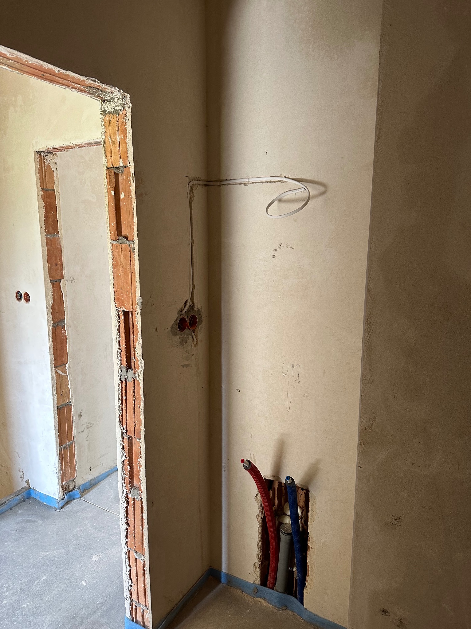 Wnętrze w trakcie remontu: widoczne przewody elektryczne wystające ze ściany, rury wodne w bruździe oraz surowa ościeżnica drzwiowa z cegły, zabezpieczona folią na podłodze.