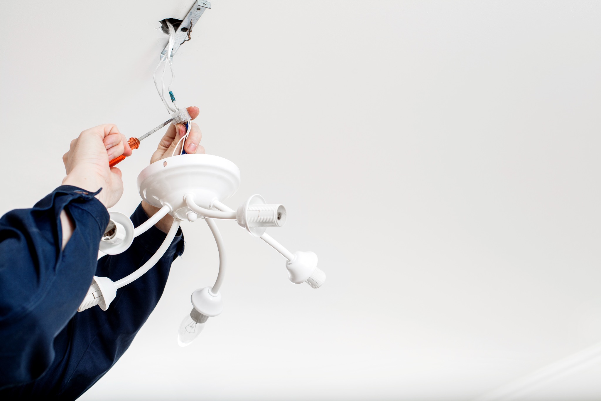 Elektryk w niebieskiej koszuli montuje białą, nowoczesną lampę sufitową z trzema ramionami i żarówką, używając pomarańczowego śrubokręta do połączenia przewodów.