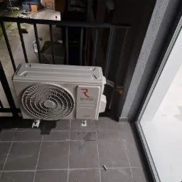 Jednostka zewnętrzna klimatyzatora Rotenso zamontowana na balkonie, widok z góry, szare płytki podłogowe, fragment balustrady i okna balkonowego.