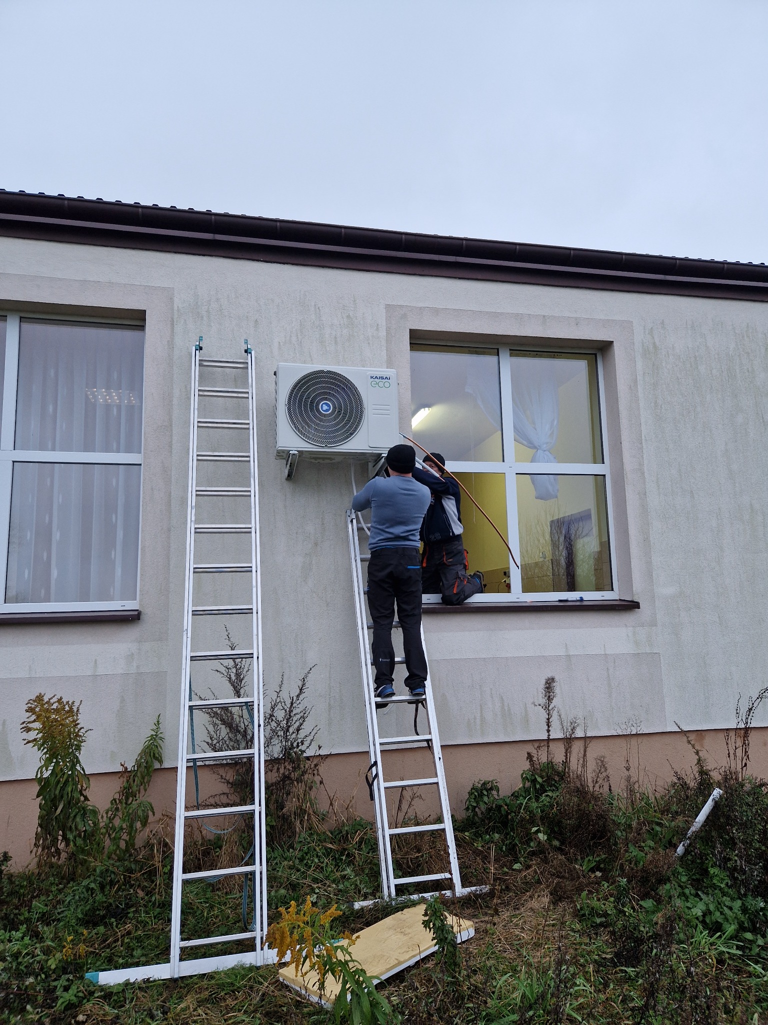 Dwóch monterów na drabinach instaluje zewnętrzną jednostkę klimatyzacji KAIHAI na ścianie budynku z dwoma oknami, widoczne narzędzia i przewody.