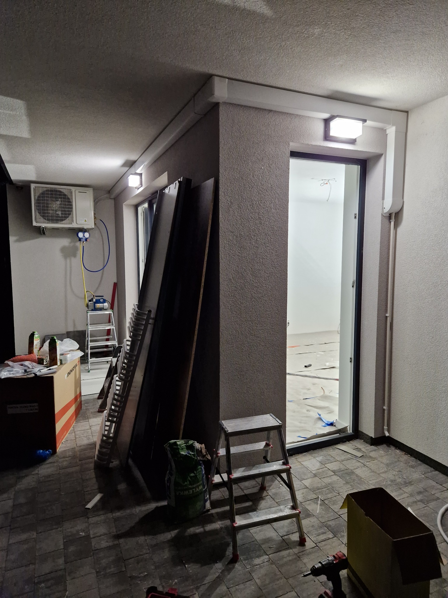 Wnętrze w trakcie montażu klimatyzacji: jednostka zewnętrzna zamontowana na ścianie, podłączona do pompy próżniowej za pomocą węży, drabina, narzędzia, płyty i worek z zaprawą na podłodze z płytek.