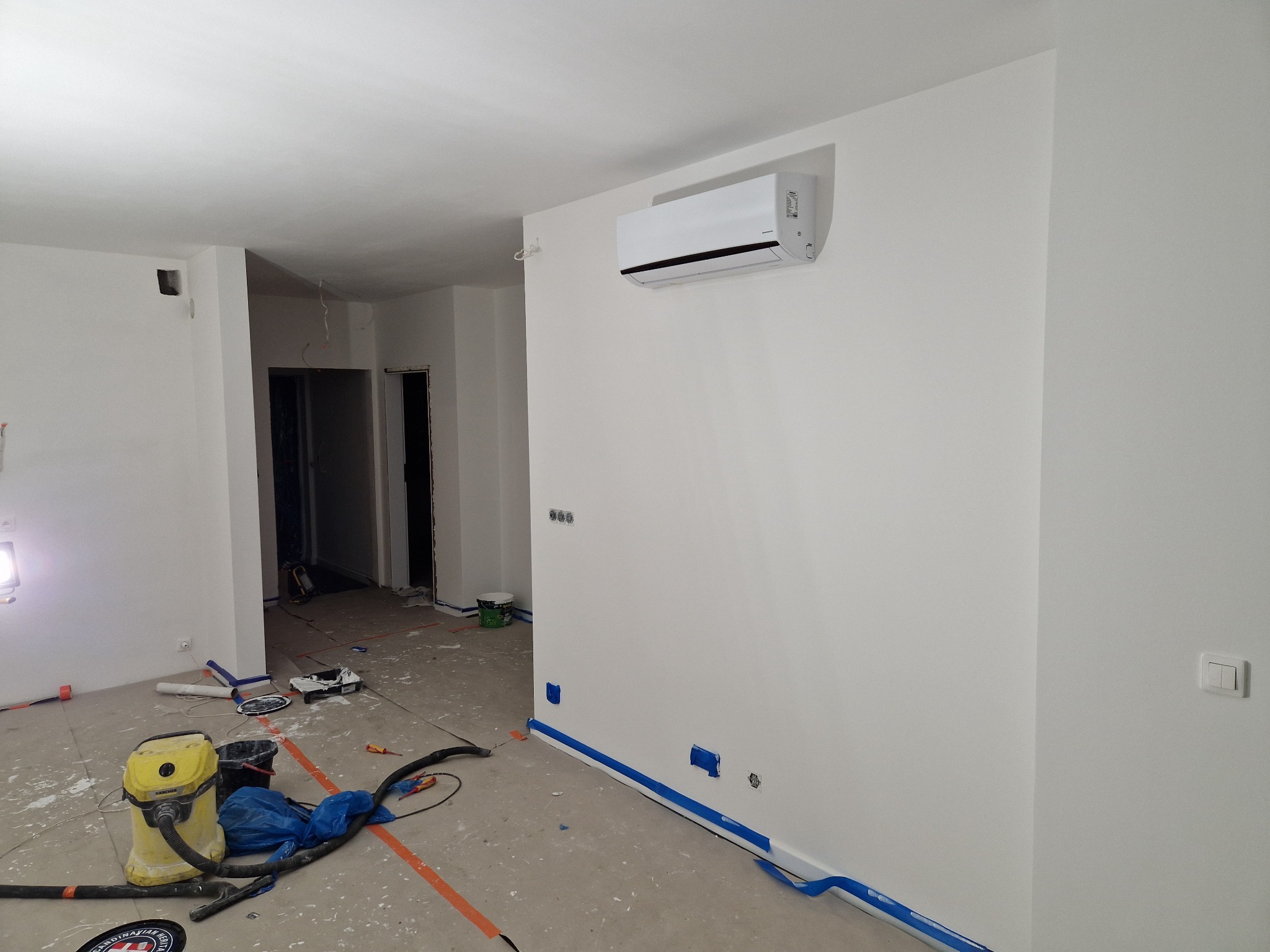 Biały klimatyzator ścienny zamontowany w świeżo wyremontowanym, pustym pokoju z widocznymi śladami po remoncie, takimi jak taśmy malarskie, plamy farby i żółty odkurzacz przemysłowy Karcher.