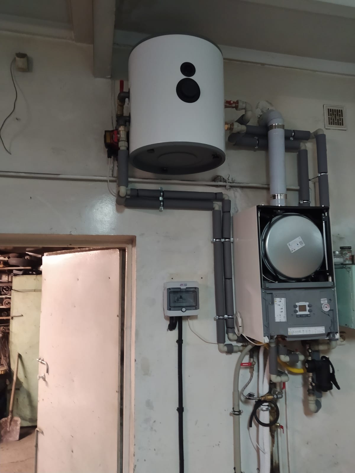 Nowoczesny, biały bojler elektryczny z szarym akcentem, zamontowany nad otwartym piecem gazowym w kotłowni z widoczną instalacją hydrauliczną w szarych otulinach i otwartymi drzwiami...