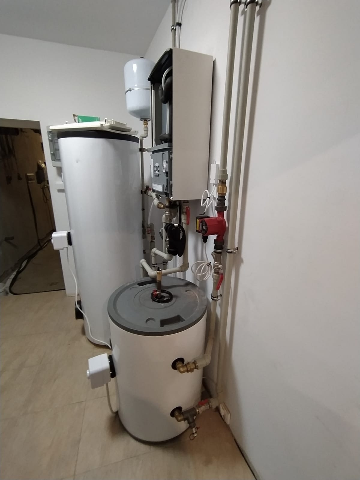 Kocioł gazowy kondensacyjny z podłączeniami hydraulicznymi, widoczny również zasobnik ciepłej wody i pompa obiegowa, instalacja w pomieszczeniu technicznym.