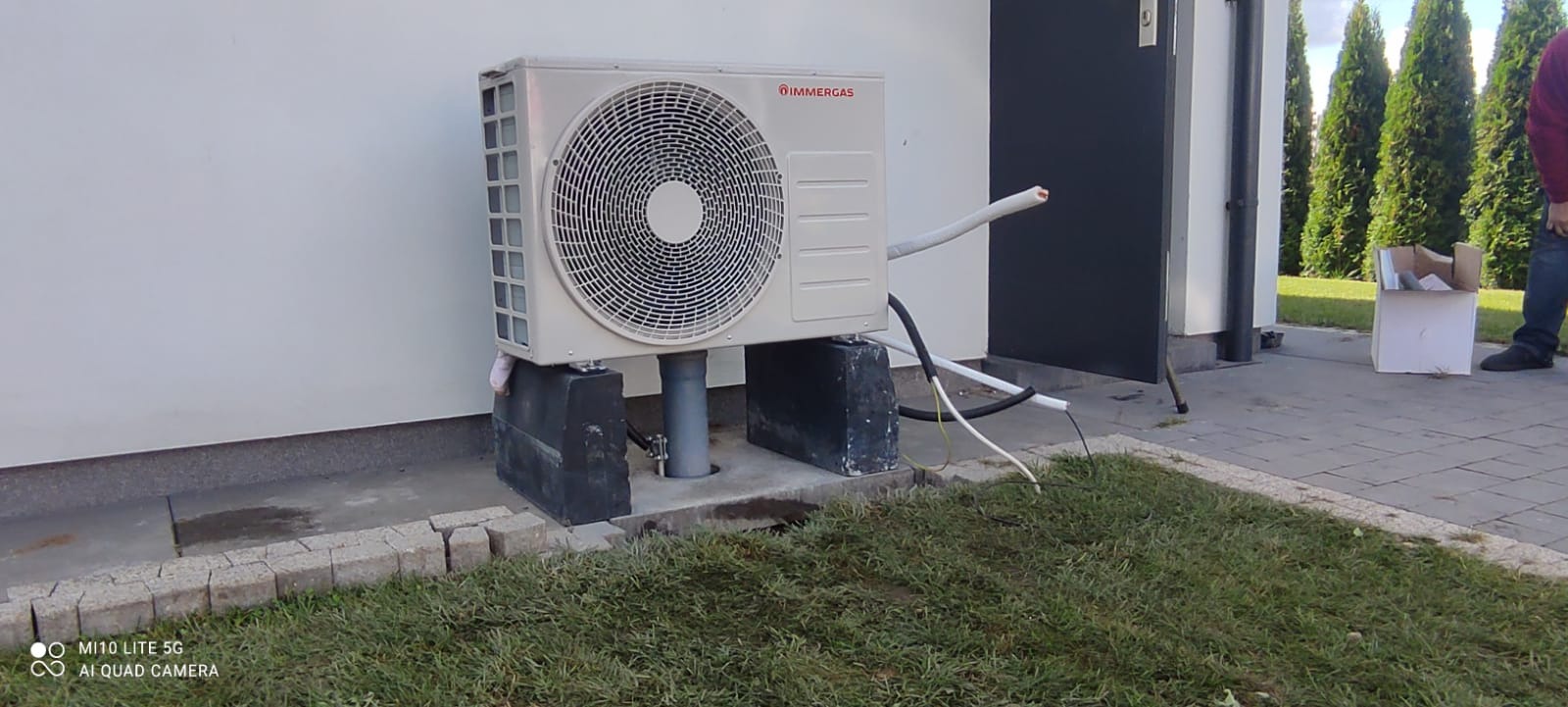 Zewnętrzna jednostka klimatyzacji Immergas zamontowana na betonowych blokach przy białej ścianie budynku, widoczne przewody i rury instalacyjne, obok drzwi i trawnik.