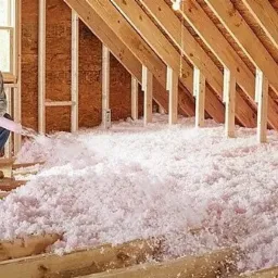 Pracownik w masce i okularach ochronnych aplikuje różową izolację z długiej rury w przestrzeni poddasza z widocznymi drewnianymi belkami stropowymi i oknem w tle.