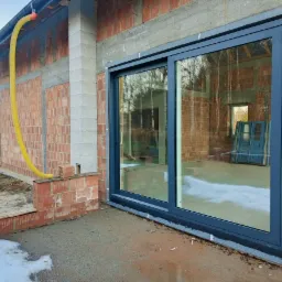 Surowe wykończenie domu w trakcie budowy z zamontowanym dużym, przesuwnym oknem z ciemną ramą; widoczny mur z czerwonej cegły, żółty wąż wentylacyjny i częściowo ośnieżony teren.