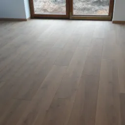 Ułożona podłoga panelowa w odcieniach brązu, widok z perspektywy, w tle okno z widokiem na ziemię i roślinność.