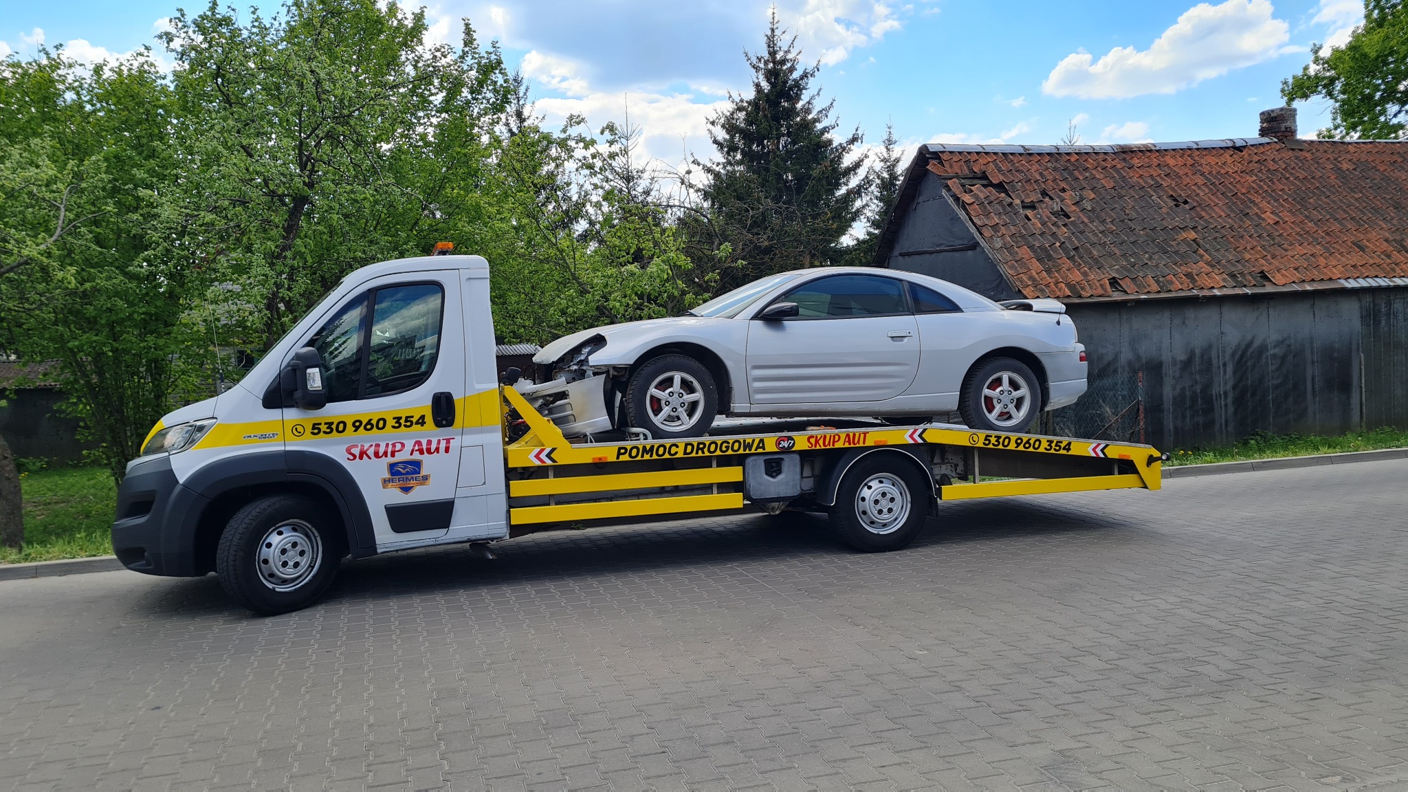 Biała laweta z uszkodzonym srebrnym samochodem osobowym na platformie, widoczne numery telefonu i napis 'SKUP AUT' na kabinie, w tle budynek z zniszczonym dachem.