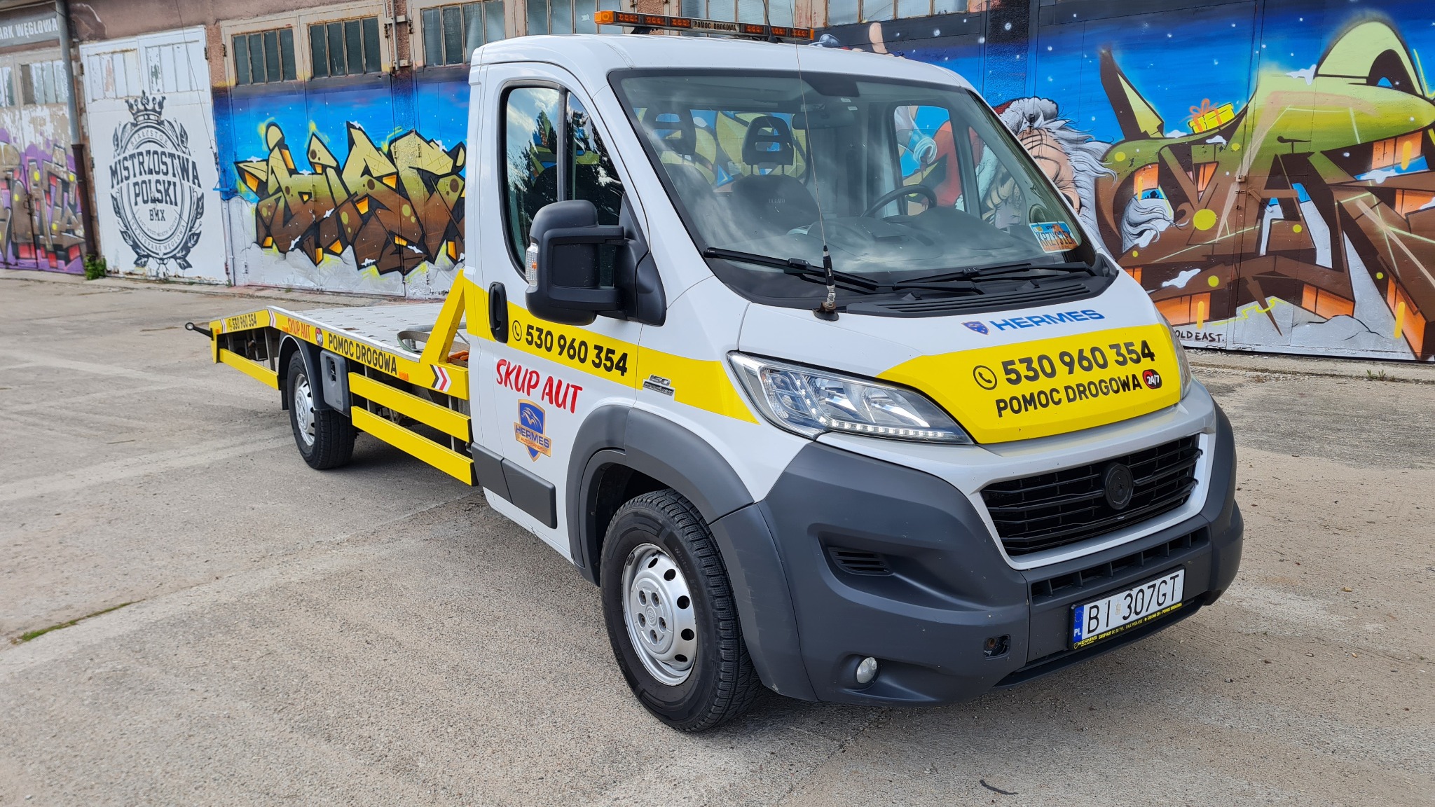 Biała laweta z żółtymi akcentami i numerem telefonu na tle betonowego placu i graffiti na ścianach budynków.