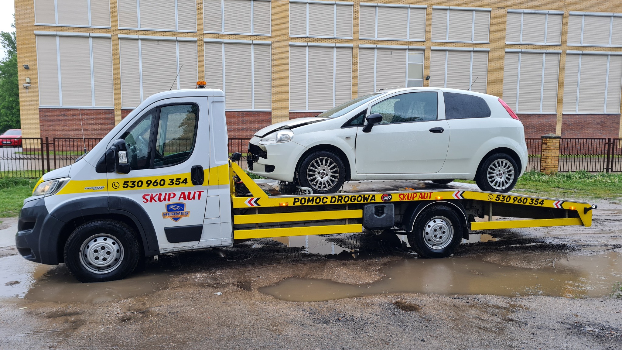 Biała laweta z uszkodzonym białym Fiatem Punto na platformie, zaparkowana na mokrym asfalcie przed budynkiem z cegły