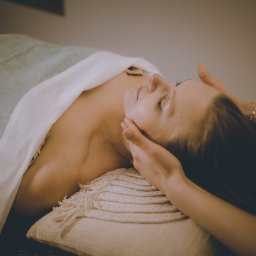 Bakasana Julia Jakubowicz - Klientka z zamkniętymi oczami podczas masażu twarzy i dekoltu, leżąca na miękkim podłożu z dekoracyjną poduszką i ręcznikiem.