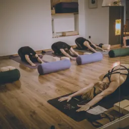 Grupa osób ćwiczy pozycję dziecka na matach do ćwiczeń w studiu, widoczne bloki do jogi i wałki, podłoga z paneli, stonowane oświetlenie.