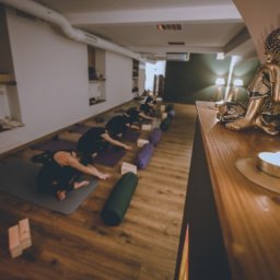 Bakasana Julia Jakubowicz - Grupa osób wykonuje pozycję dziecka na matach w studiu z drewnianą podłogą; w tle posąg Buddy i zapalona świeca na drewnianym blacie.