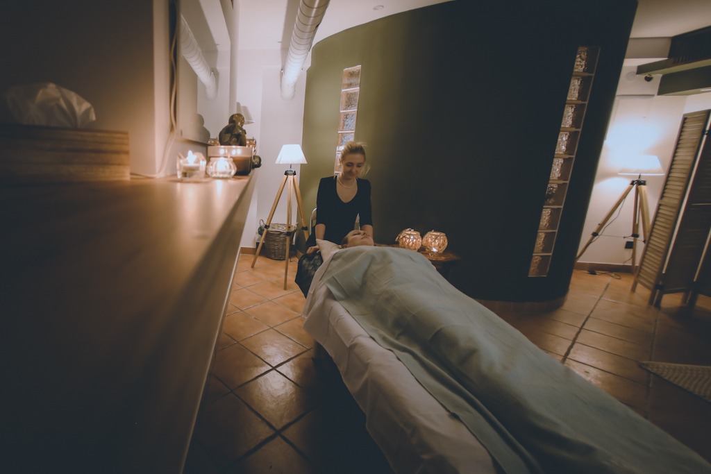 Kobieta w czarnym stroju wykonuje masaż głowy osobie leżącej na stole w gabinecie z terakotową podłogą, oświetlonym lampami na trójnogach i świecami.