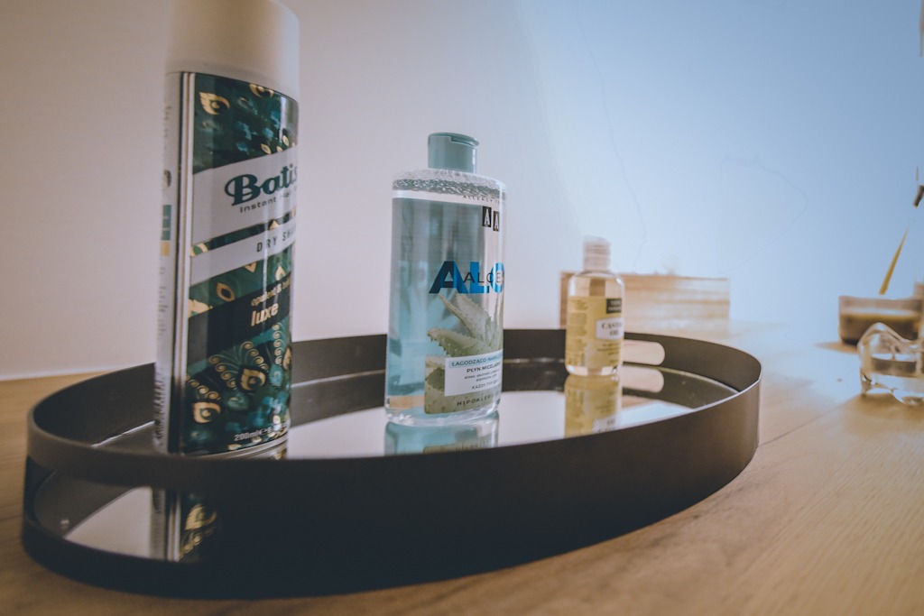 Zestaw kosmetyków: suchy szampon Batiste, płyn micelarny Aloes i olejek rycynowy, ułożone na okrągłej, czarnej tacy z lustrzanym dnem na drewnianym blacie.