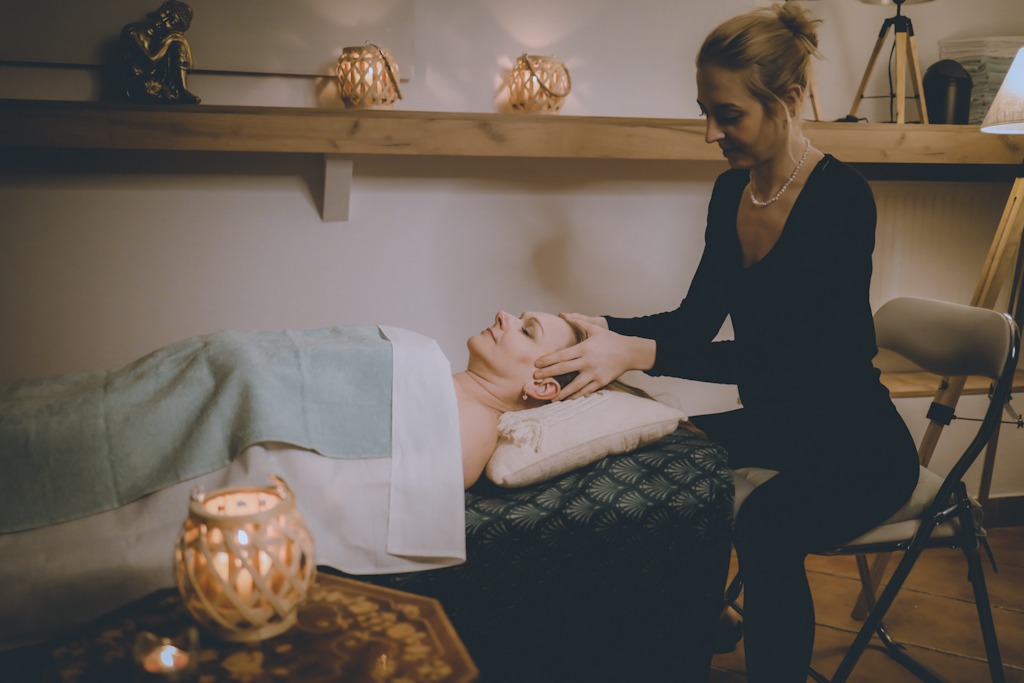 Kobieta w czarnym stroju wykonuje masaż głowy klientki leżącej na stole przykrytym turkusowym pledem, w tle półka z dekoracyjnymi lampionami.