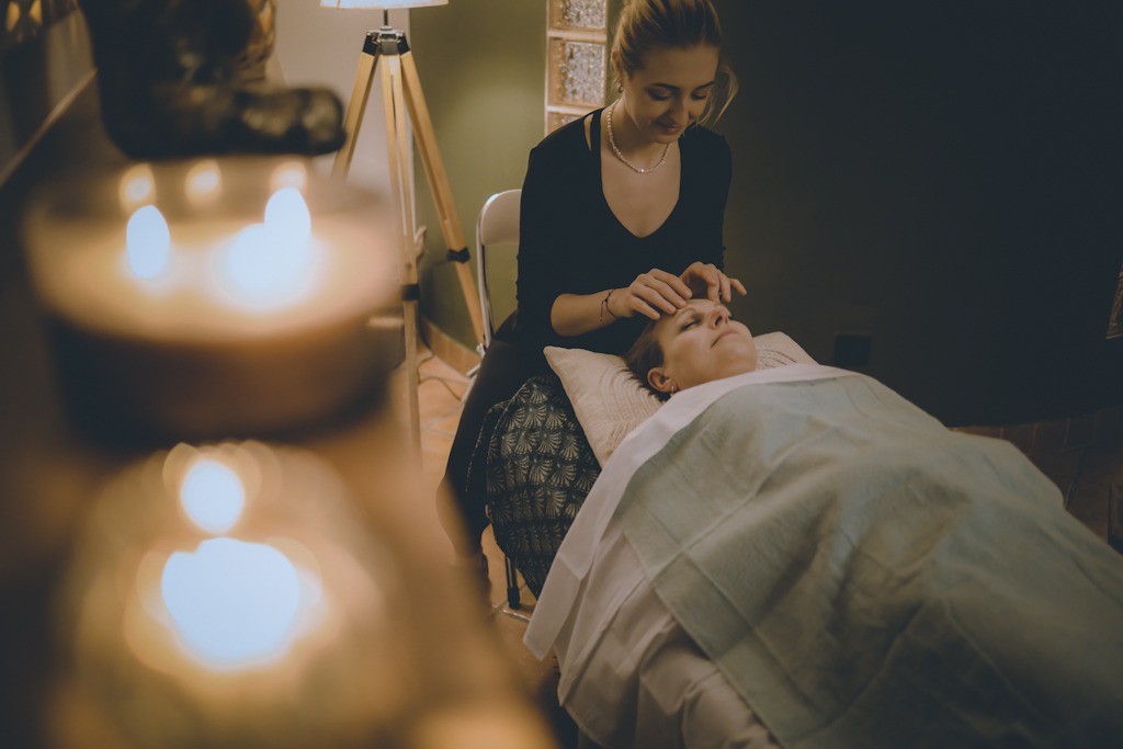 Kobieta otrzymuje masaż głowy w gabinecie, z rozproszonym światłem świec w tle i relaksującą atmosferą.