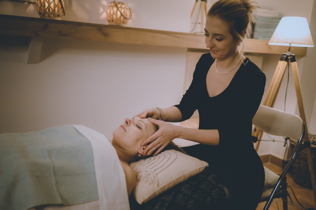 Kobieta w czarnej sukience wykonuje masaż twarzy klientce leżącej na stole do masażu z beżową poduszką i turkusowym okryciem, w tle drewniana półka z dekoracyjnymi lampionami i lampa na trójnogu.