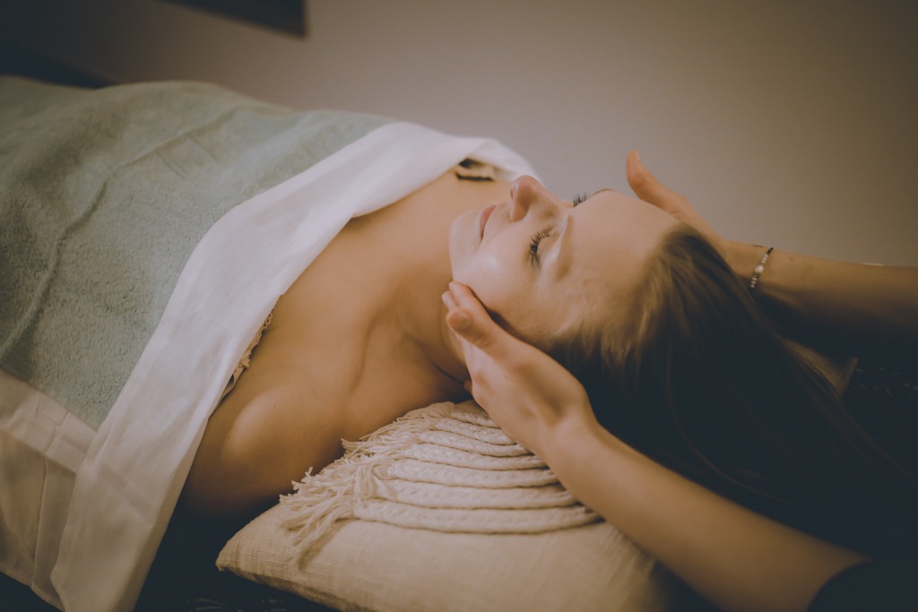 Klientka z zamkniętymi oczami podczas masażu twarzy i dekoltu, leżąca na miękkim podłożu z dekoracyjną poduszką i ręcznikiem.