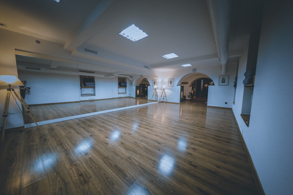 Jasne, przestronne studio z drewnianą podłogą, lustrami na ścianie i oświetleniem sufitowym. Widoczne lampy na drewnianych trójnogach oraz dekoracje w niszach ściennych.