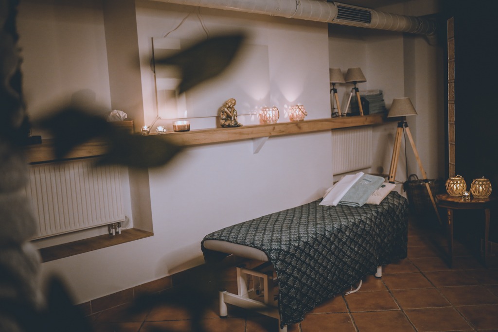 Przytulne wnętrze gabinetu masażu z łóżkiem do masażu przykrytym wzorzystą narzutą, dekoracyjną półką ze świecami i figurką, oraz lampami na drewnianych trójnogach, oświetlonym ciepłym światłem.