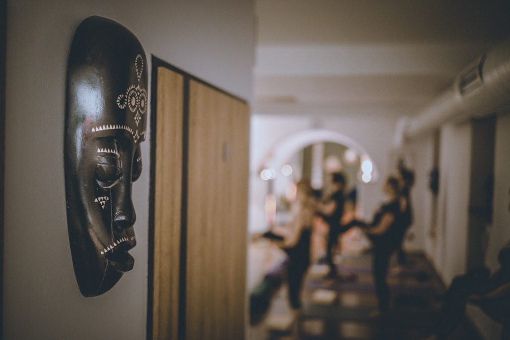 Afrykańska maska na ścianie w studiu, w tle grupa osób ćwiczących w pozycji stojącej na matach.