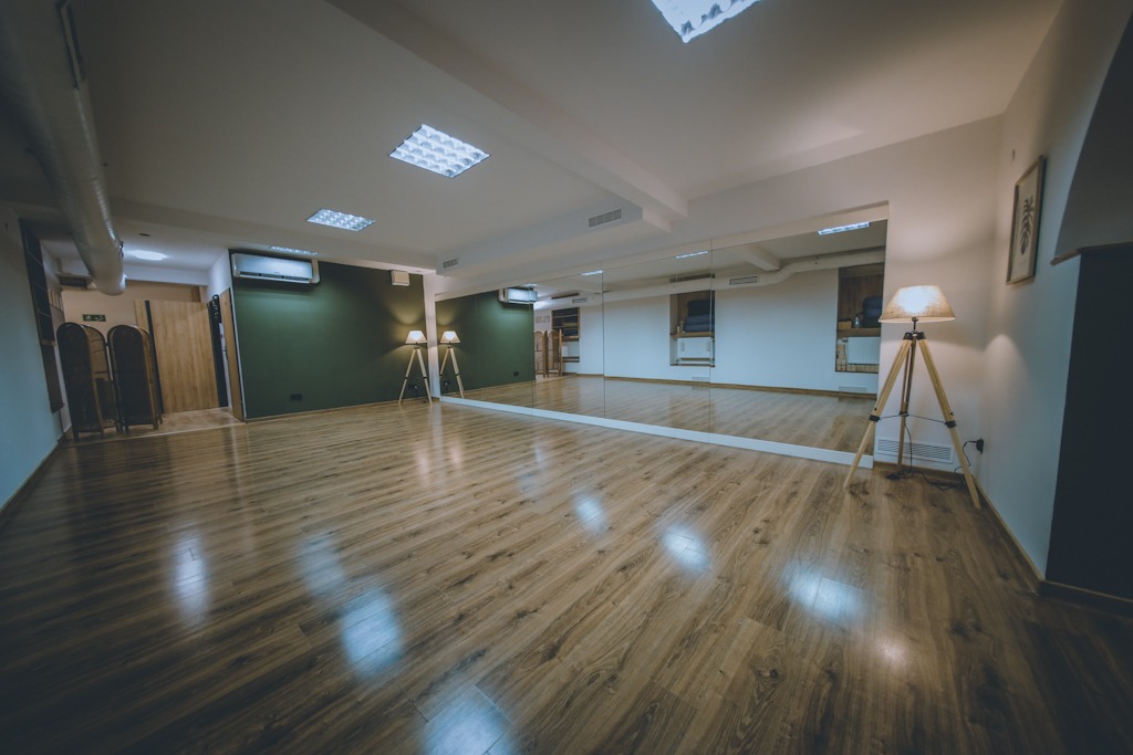 Pusta sala do ćwiczeń z drewnianą podłogą, dużym lustrem na ścianie, dwoma lampami trójnożnymi i klimatyzacją, ściana w kolorze butelkowej zieleni.