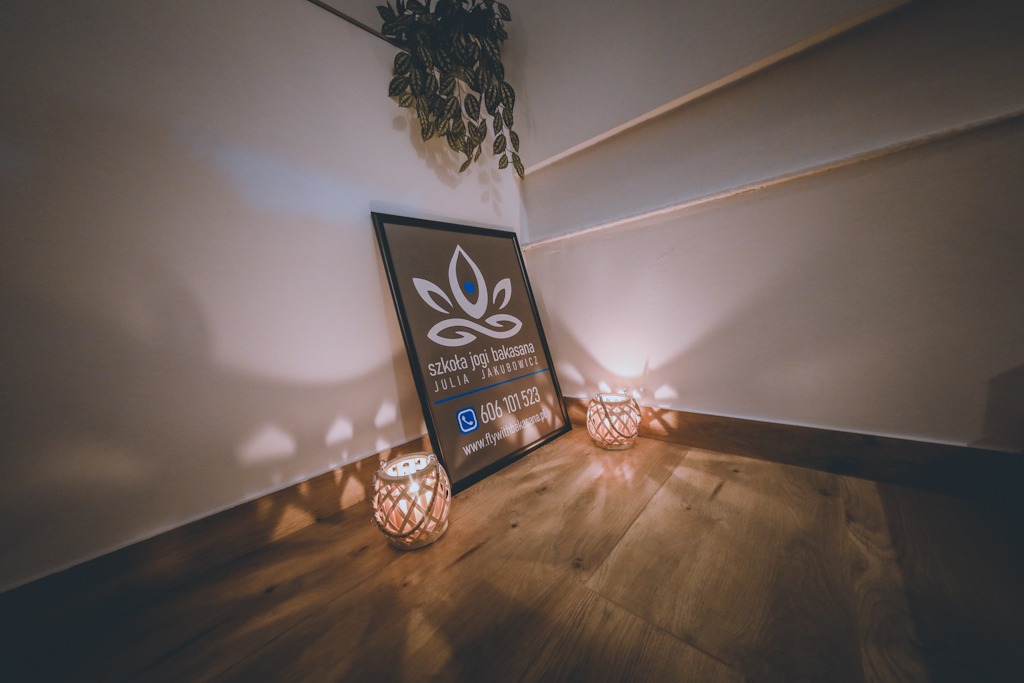Kącik z plakatem szkoły jogi bakasana Julia Jakubowicz, oświetlony dwoma lampionami ze świecami, wisząca roślina na ścianie, drewniana podłoga.