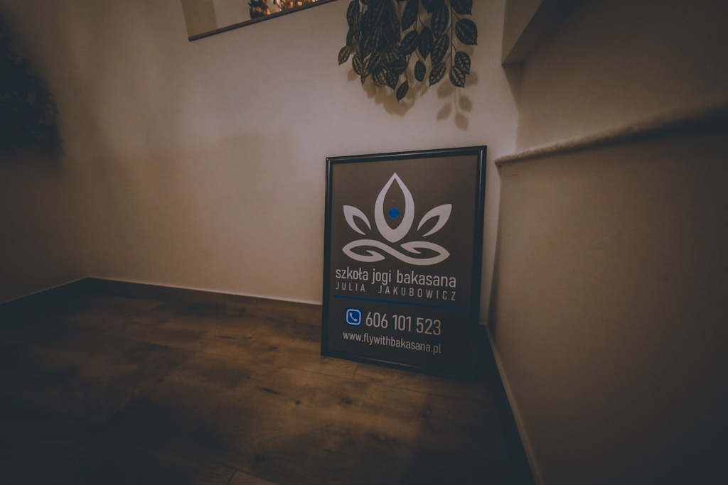 Tablica informacyjna 'Szkoła Jogi Bakasana Julia Jakubowicz' z numerem telefonu i adresem strony internetowej, umieszczona w rogu pomieszczenia na drewnianej podłodze w Rzeszowie, z rośliną...