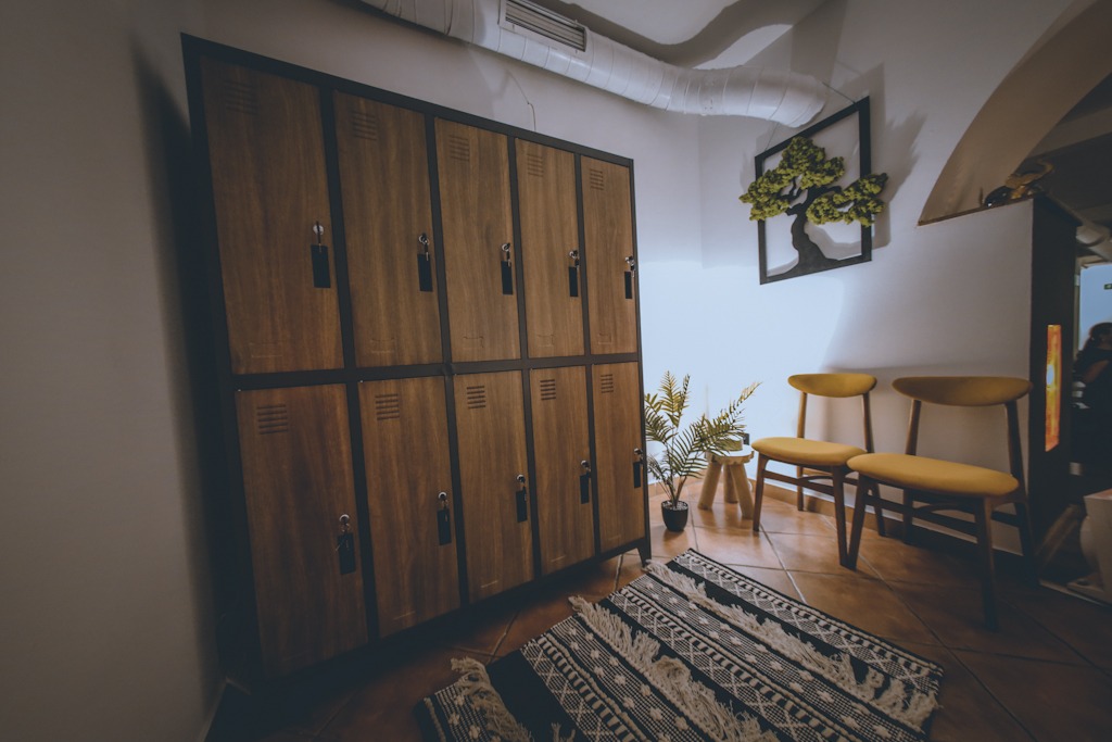 Szereg drewnianych szafek z czarnymi obramowaniami w szatni studia jogi, obok dwie żółte krzesła, dekoracja ścienna w kształcie drzewa bonsai i dywanik z frędzlami.