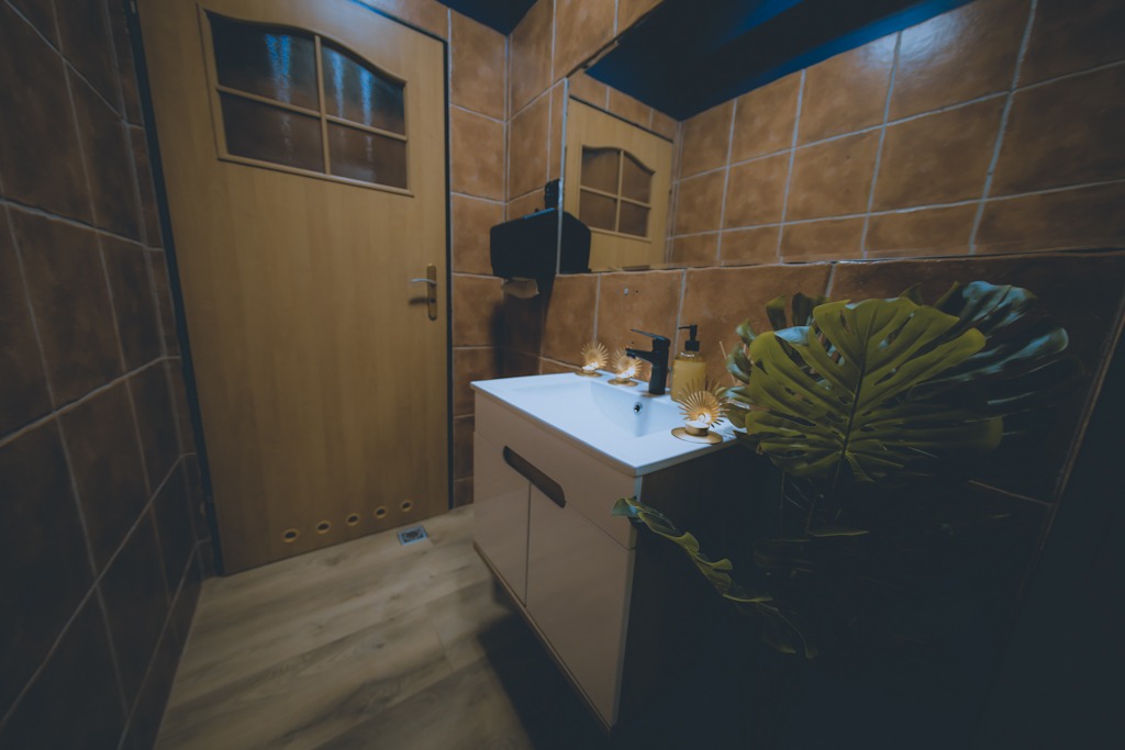 Umywalka z białym blatem i szafką, armatura w kolorze czarnym oraz dodatki w złotym odcieniu, dekoracja roślinna z liści monstery, brązowe płytki na ścianach, drewniane drzwi z szybami.