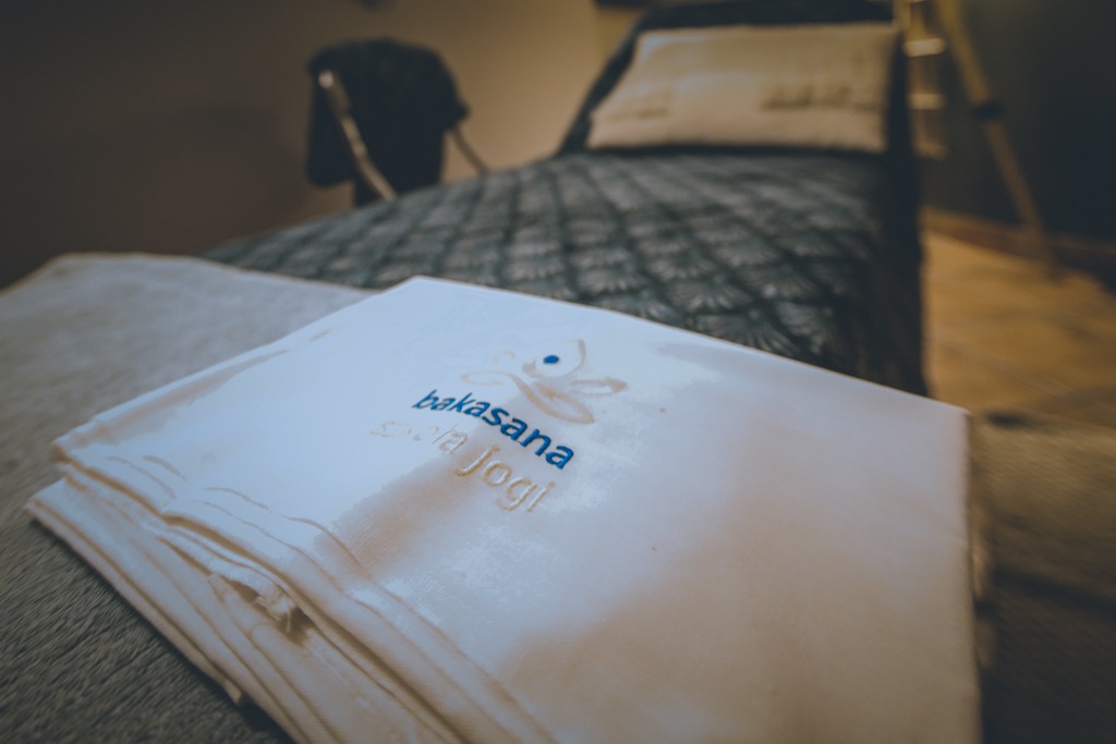 Stos białych ręczników z wyhaftowanym logo 'bakasana szkoła jogi' leży na stole w tle łóżko i drabina.