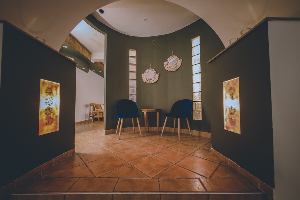 Wnętrze poczekalni z dwoma granatowymi krzesłami i małym stolikiem, dekoracjami ściennymi z makramy i podświetlanymi panelami z motywem spirali, w tle widoczne kolejne pomieszczenie.