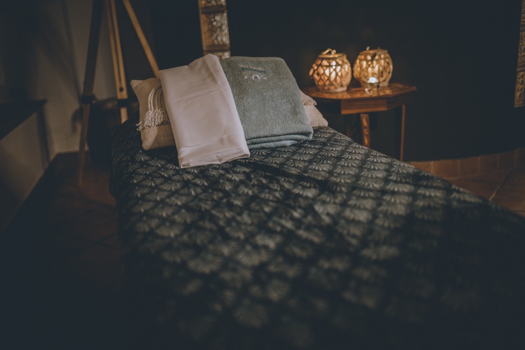 Przygotowane łóżko do masażu z ręcznikami i dekoracyjnymi lampionami na stoliku w tle, w zaciemnionym pomieszczeniu.