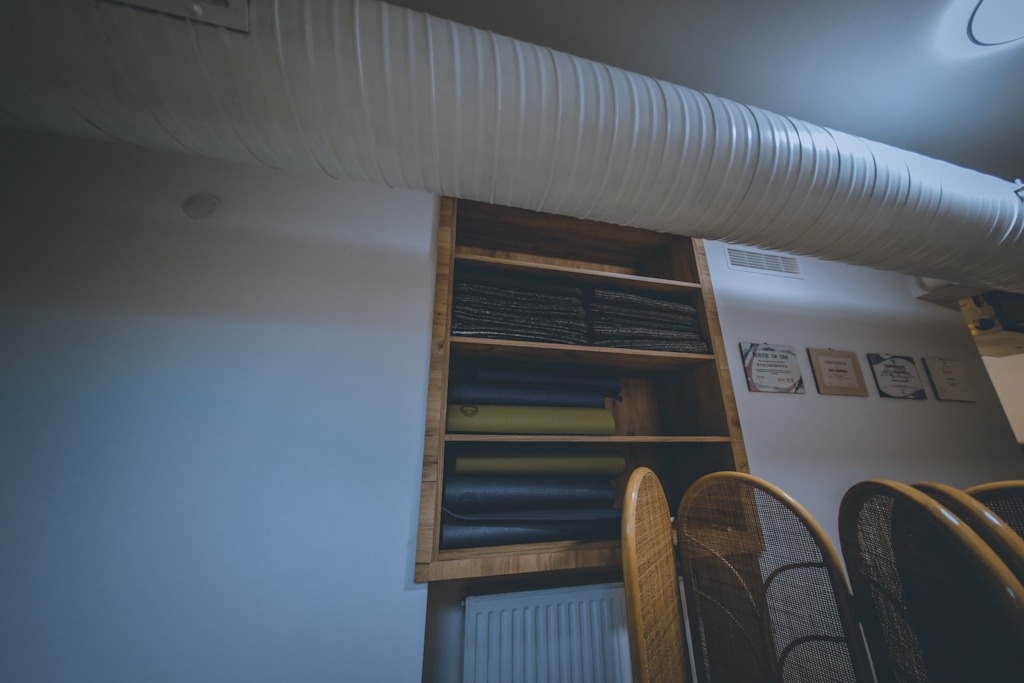 Drewniany regał z ułożonymi matami do ćwiczeń w różnych kolorach, widoczne certyfikaty na ścianie i krzesła z wikliny, biała rura wentylacyjna pod sufitem.